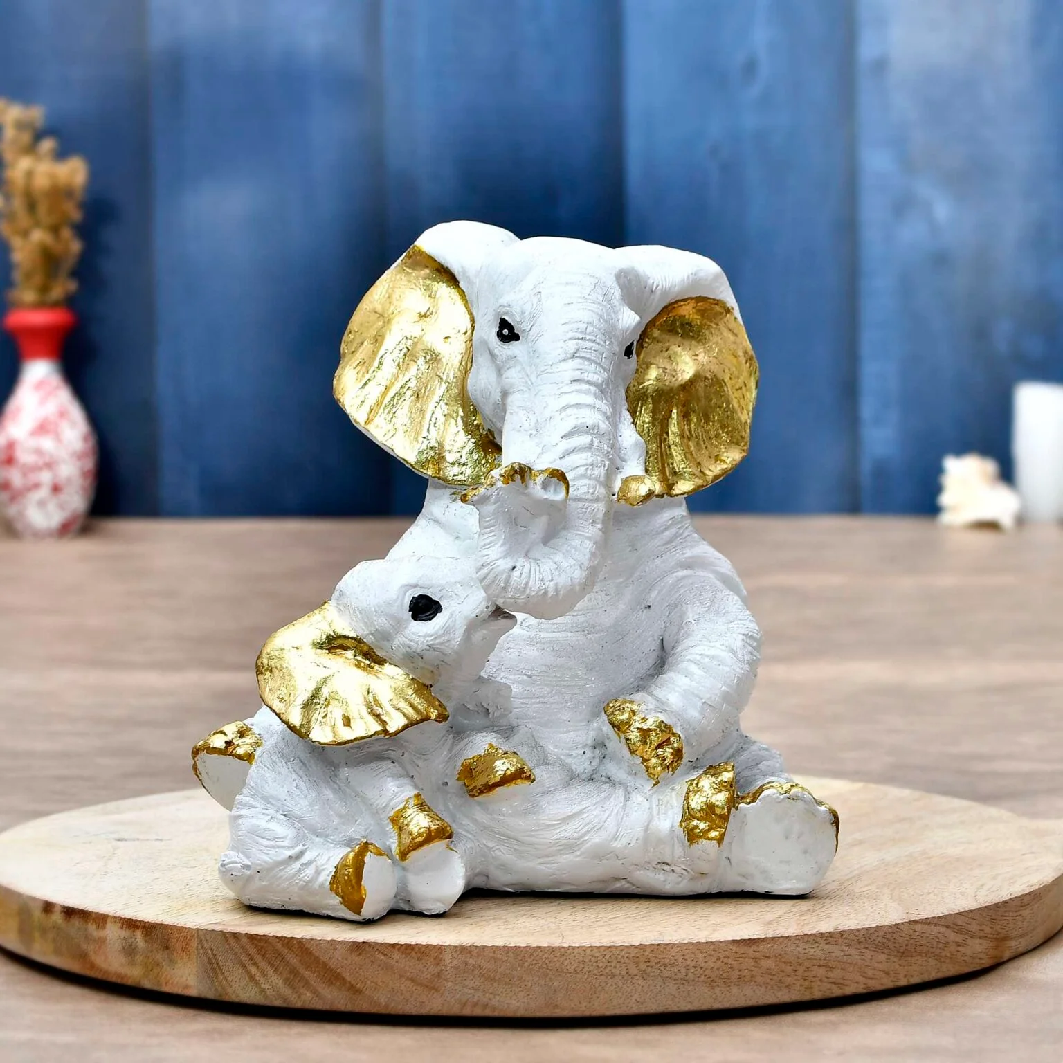 Decor Home Craft A decorative sculpture of a Polyresin Elephant to Decor Home Showpiece Figurine 29x30' multicolor R-EL1, with gold-tipped details on ears and feet, sitting on a wooden platform against a blurred blue and wooden background, perfect for enhancing the ambiance of a hotel lobby.