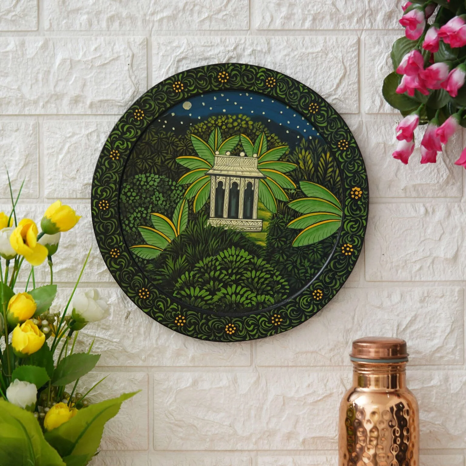 Decor Home Craft Handmade Pichwai Painting with a traditional design hanging on a white brick wall in an office, featuring a gazebo scene surrounded by lush greenery under a night sky, flanked by pink and yellow flowers and a copper vessel.