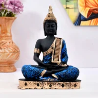 Decor Home Craft A statue of the Polyresin 27x27cm Sitting Buddha idol, adorned in a blue and gold robe, against a backdrop featuring flowers and colorful fabric, ideal for enhancing the ambiance of an office or living room.