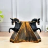 Decor Home Craft A pair of Polyresin Horse Face Antique Sculpture 17x18cm bookends in black and gold on a wooden table, with each horse rearing up against sloped, textured supports that mimic an open book. A blurred office background enhances focus on the bookends.