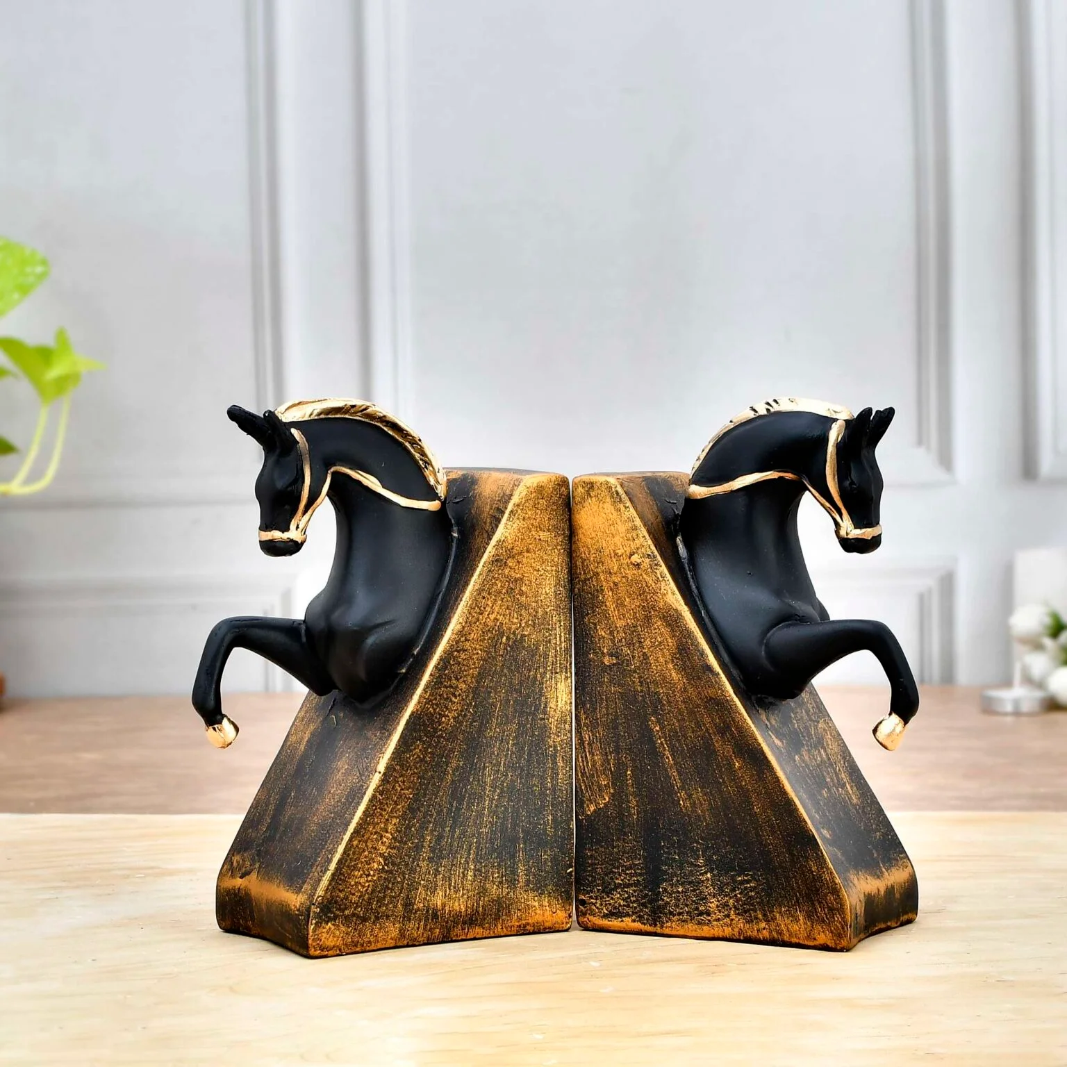 Decor Home Craft A pair of Polyresin Horse Face Antique Sculpture 17x18cm bookends in black and gold on a wooden table, with each horse rearing up against sloped, textured supports that mimic an open book. A blurred office background enhances focus on the bookends.